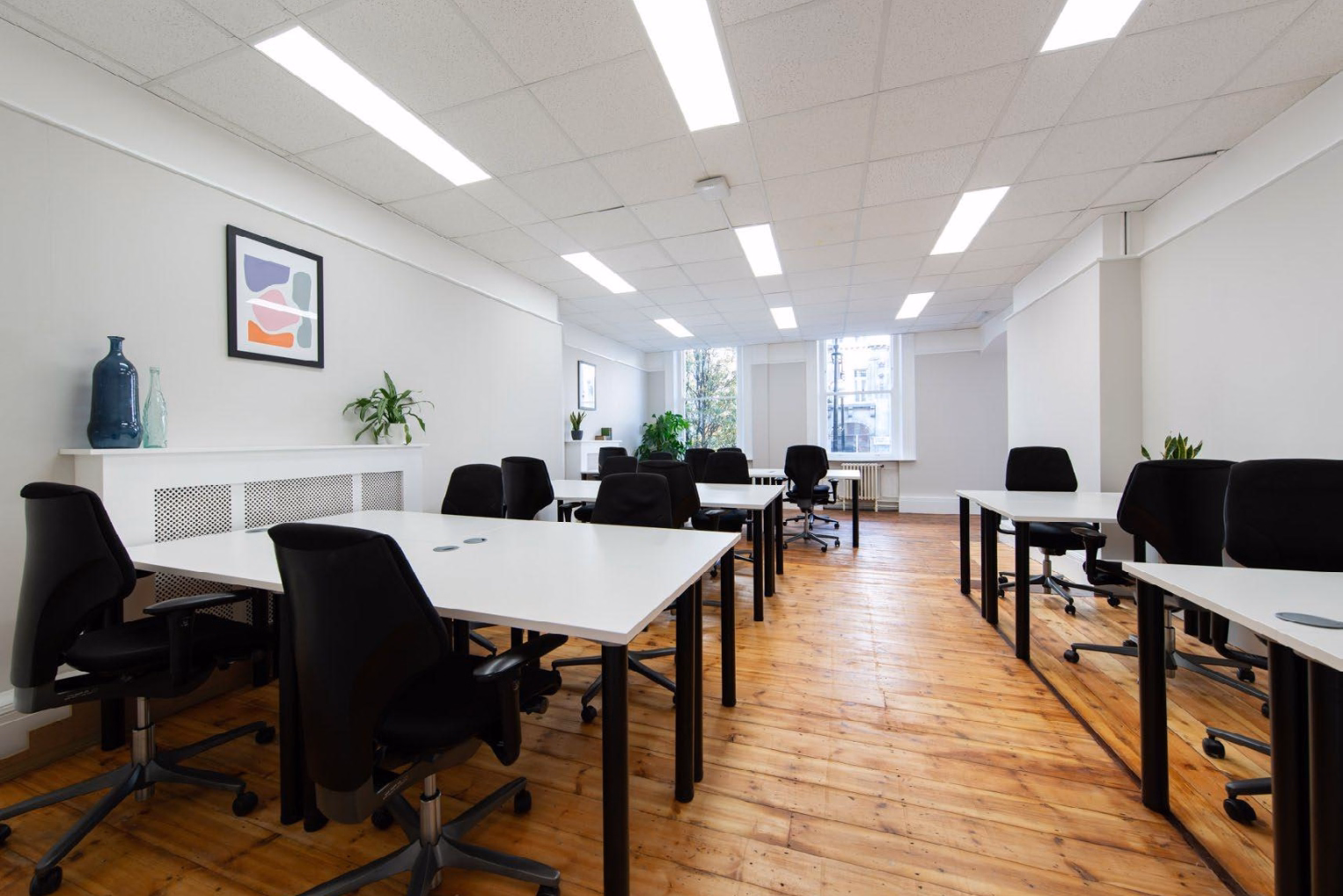 Black chairs around white desks in an open-plan workspace featuring warm hardwood floors, white walls with minimal art, and sash windows at the far end.