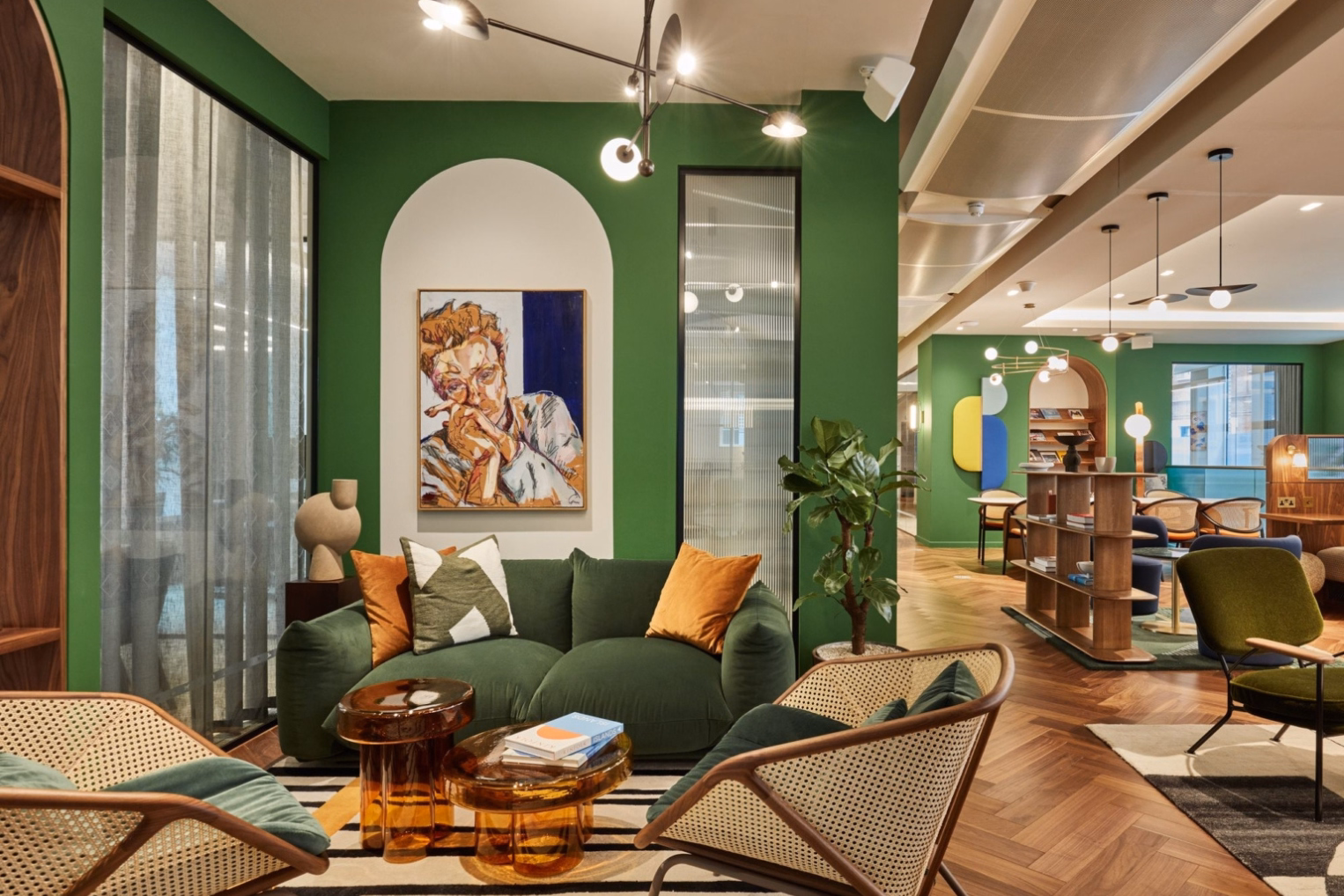 A green sofa with orange and patterned pillows with two armchairs and amber glass coffee tables near a green accent wall featuring a white arched alcove with a painting.