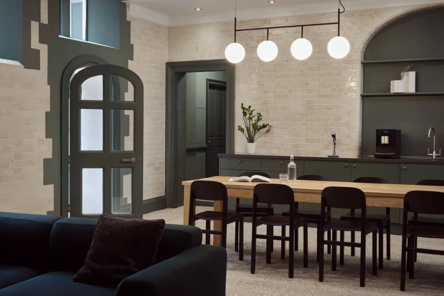 Black chairs surround a long table under a four-globe pendant light in an office kitchen and lounge area, which features olive-green cabinetry.