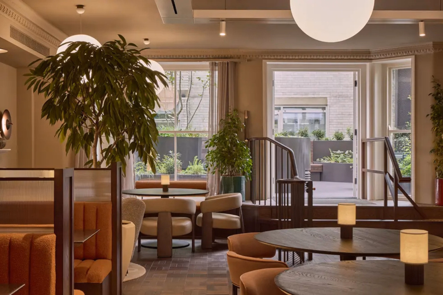 Table lamps on round wooden tables surrounded by orange and beige armchairs in a lounge, which features potted plants and glass doors leading out to the patio.