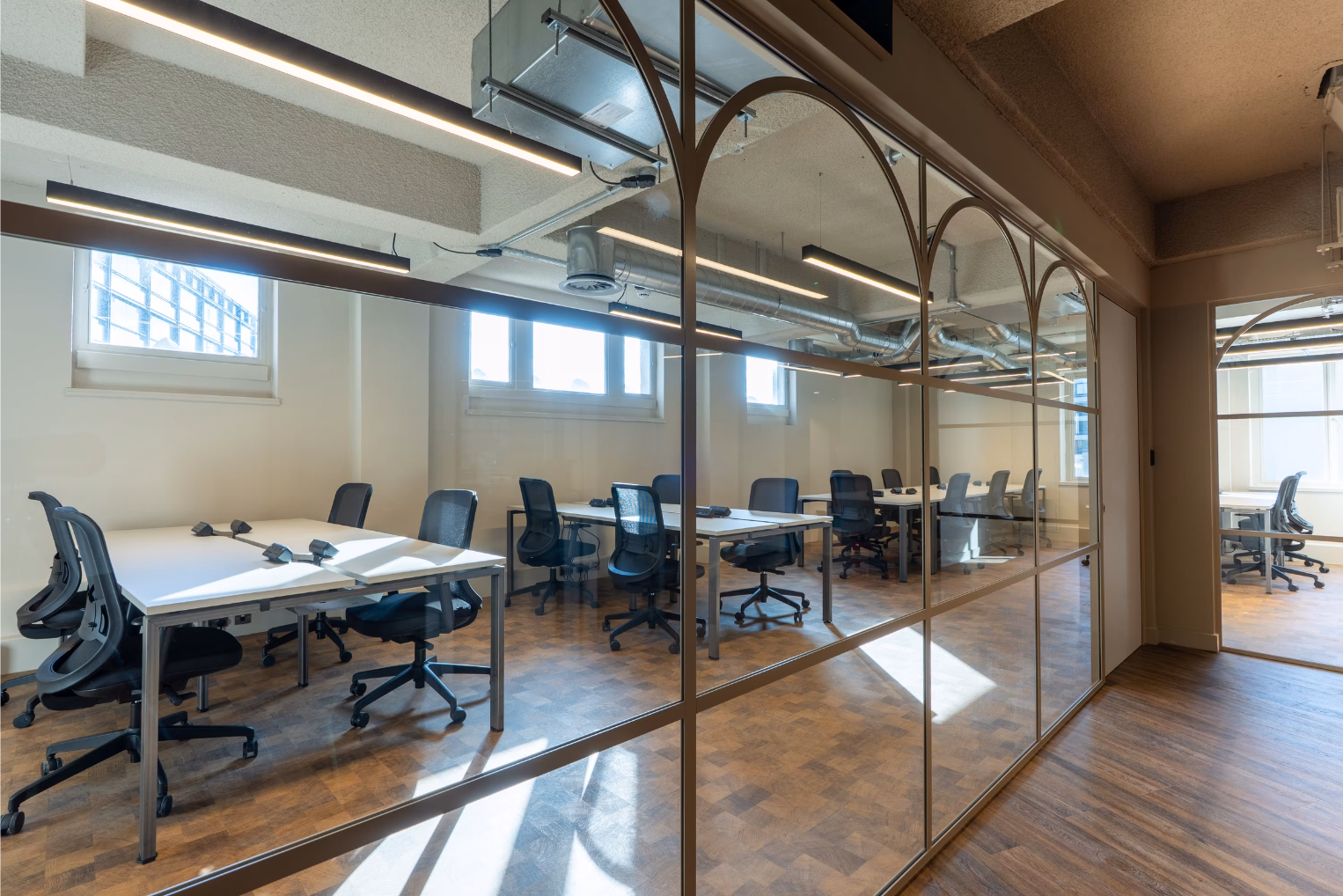 Black mesh chairs at white desks inside a bright workspace, separated by a bronze-framed glass partition wall.
