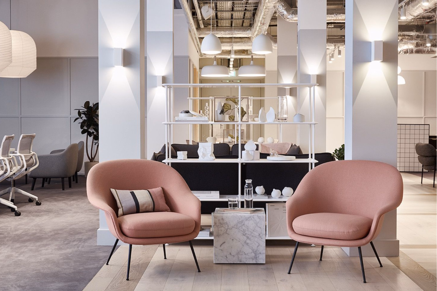 Two pink scoop-shaped armchairs on either side of a square marble side table in an open-plan office lounge, which features a white open shelving unit.