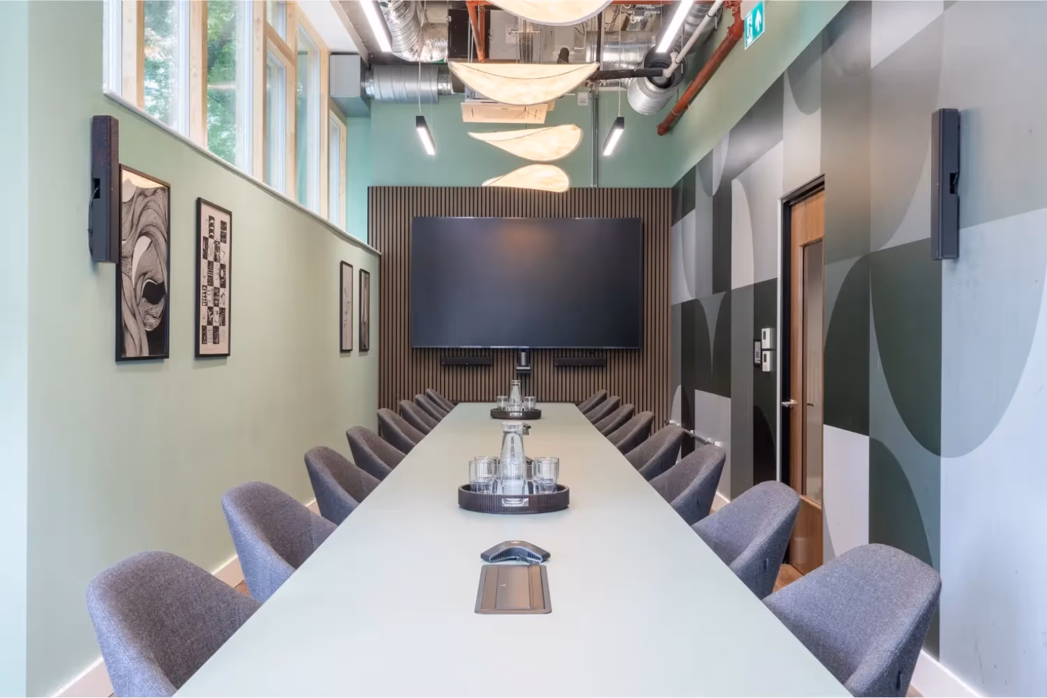 Grey armchairs surround a long table in a meeting room, which features a large wall-mounted screen against a wood-slat accent wall.