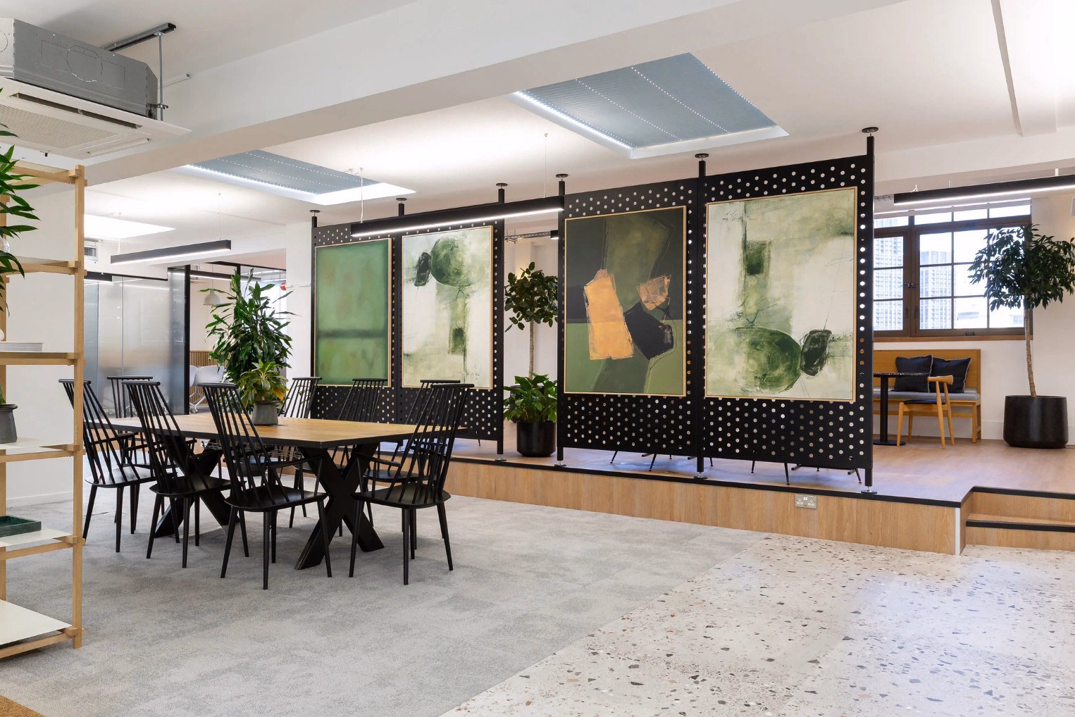 Black chairs surround a long wooden table set next to a raised wooden platform with large abstract green paintings and potted plants.