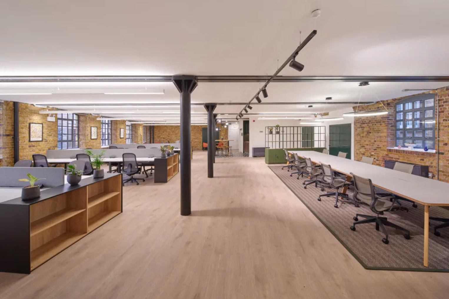 Grey chairs at individual workstations and along a long, shared hot-desking table in an open-plan office space featuring black structural pillars and exposed brick walls.