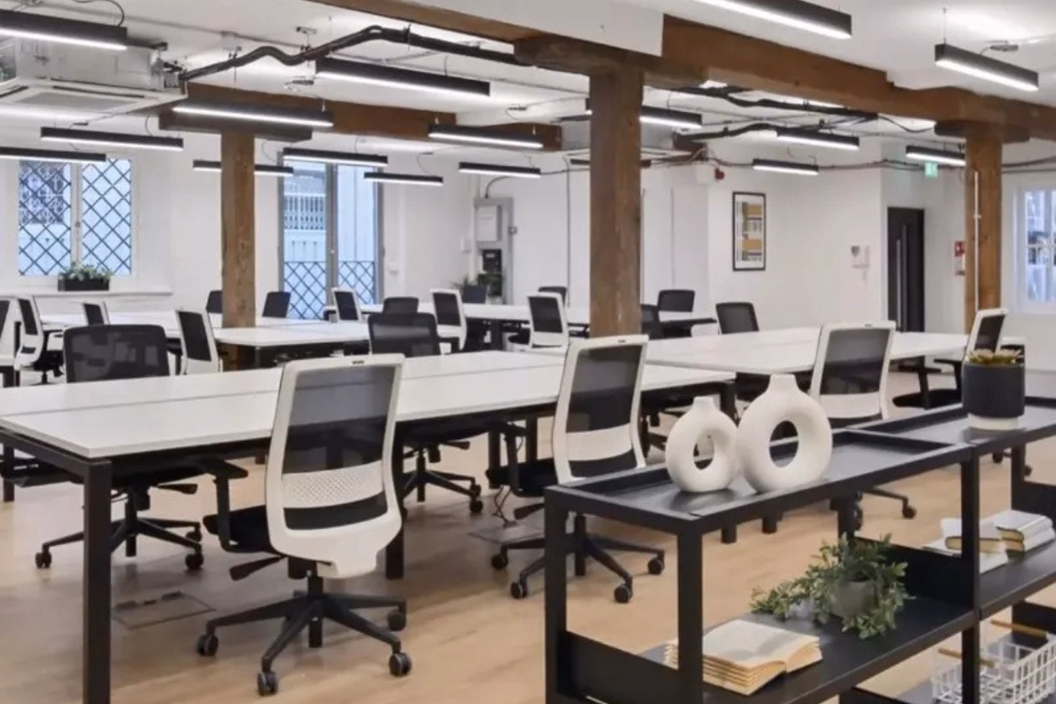 White chairs at long rows of white desks with rustic wooden support pillars, and pendant lights suspended from an industrial ceiling with exposed pipes.