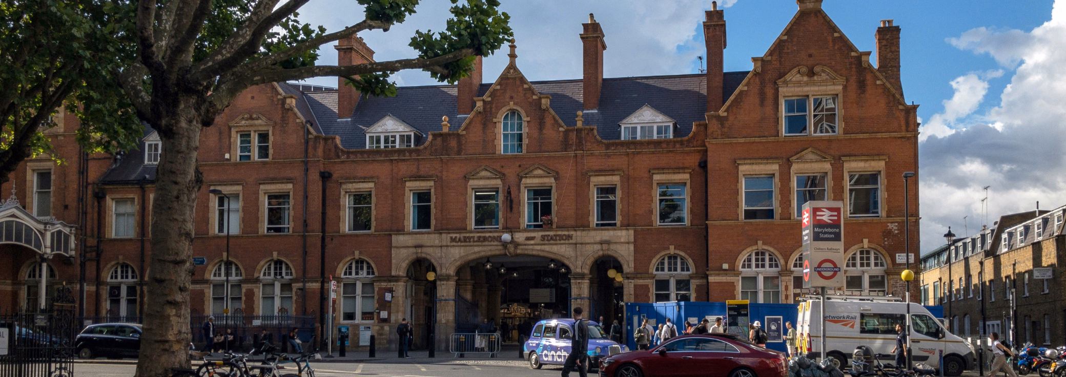 Marylebone Station features arched entrances and stone trimmings under a cloudy sky, with local traffic and parked cars.