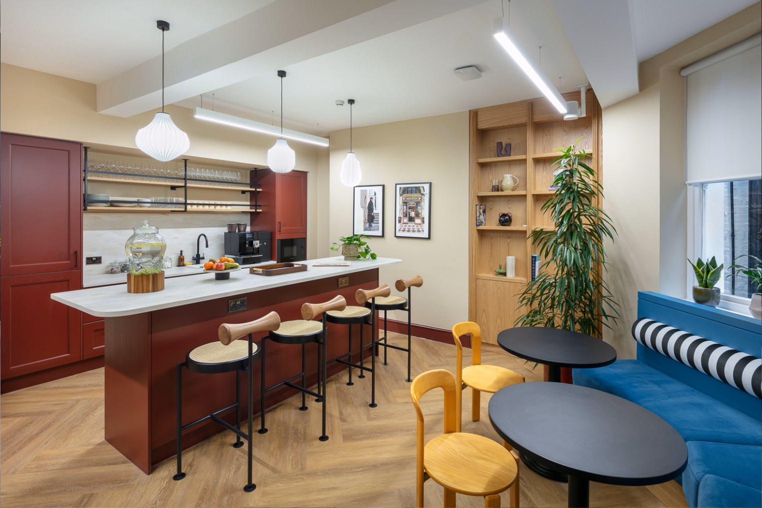 Woven-seat barstools line a kitchen island across from small black tables, yellow wooden chairs, and a blue bench with a striped pillow.