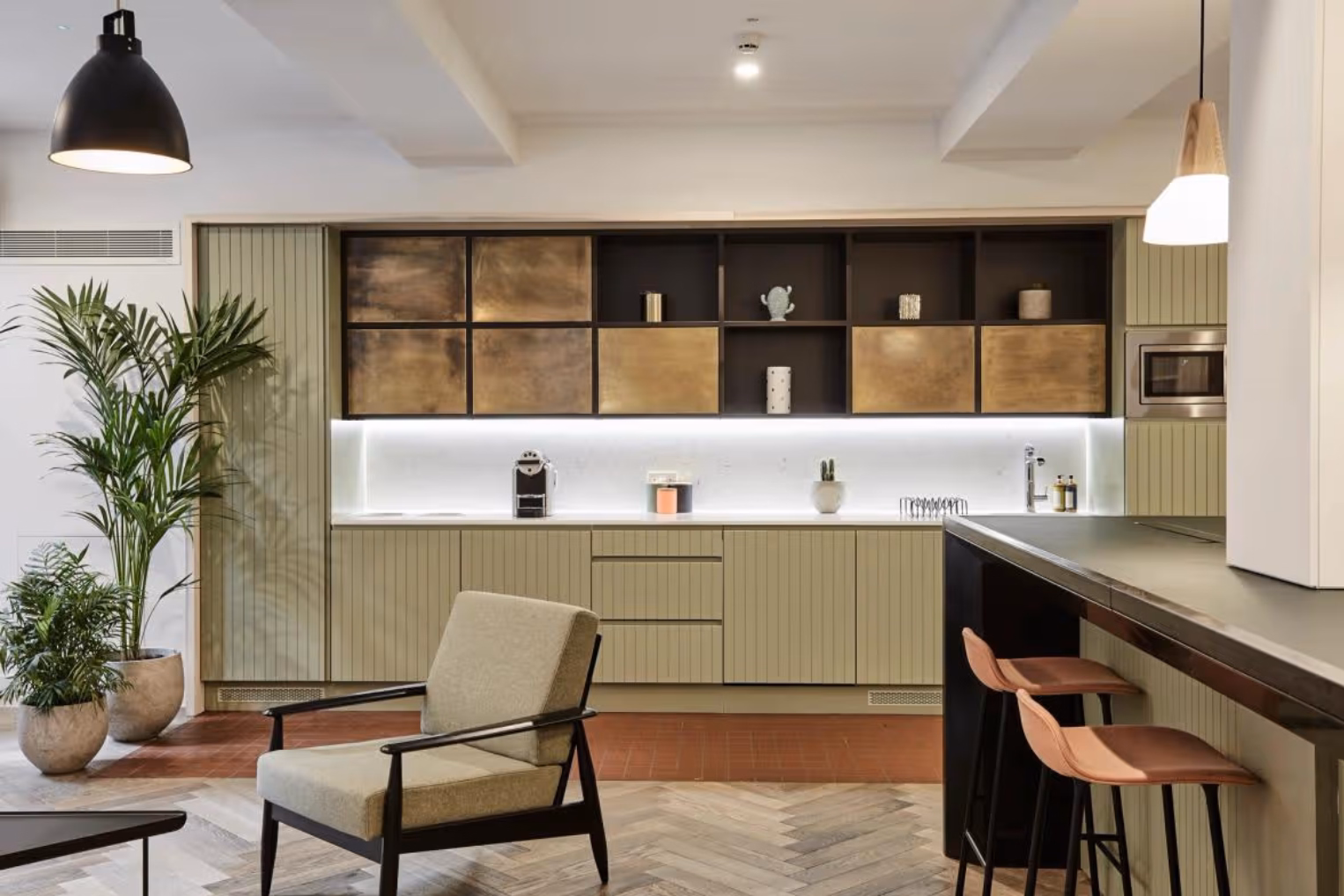 Pink barstools along a dark kitchen breakfast bar across from a light green armchair near a kitchenette with sage-green panelled cabinets.