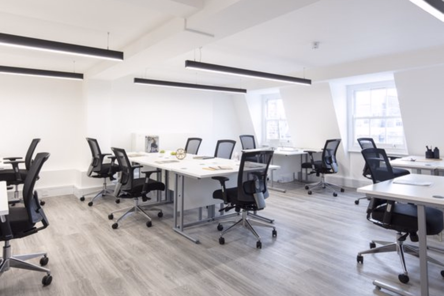 Black mesh chairs surround a long white table and separate individual desks in a bright office with white walls and black linear pendant lights.
