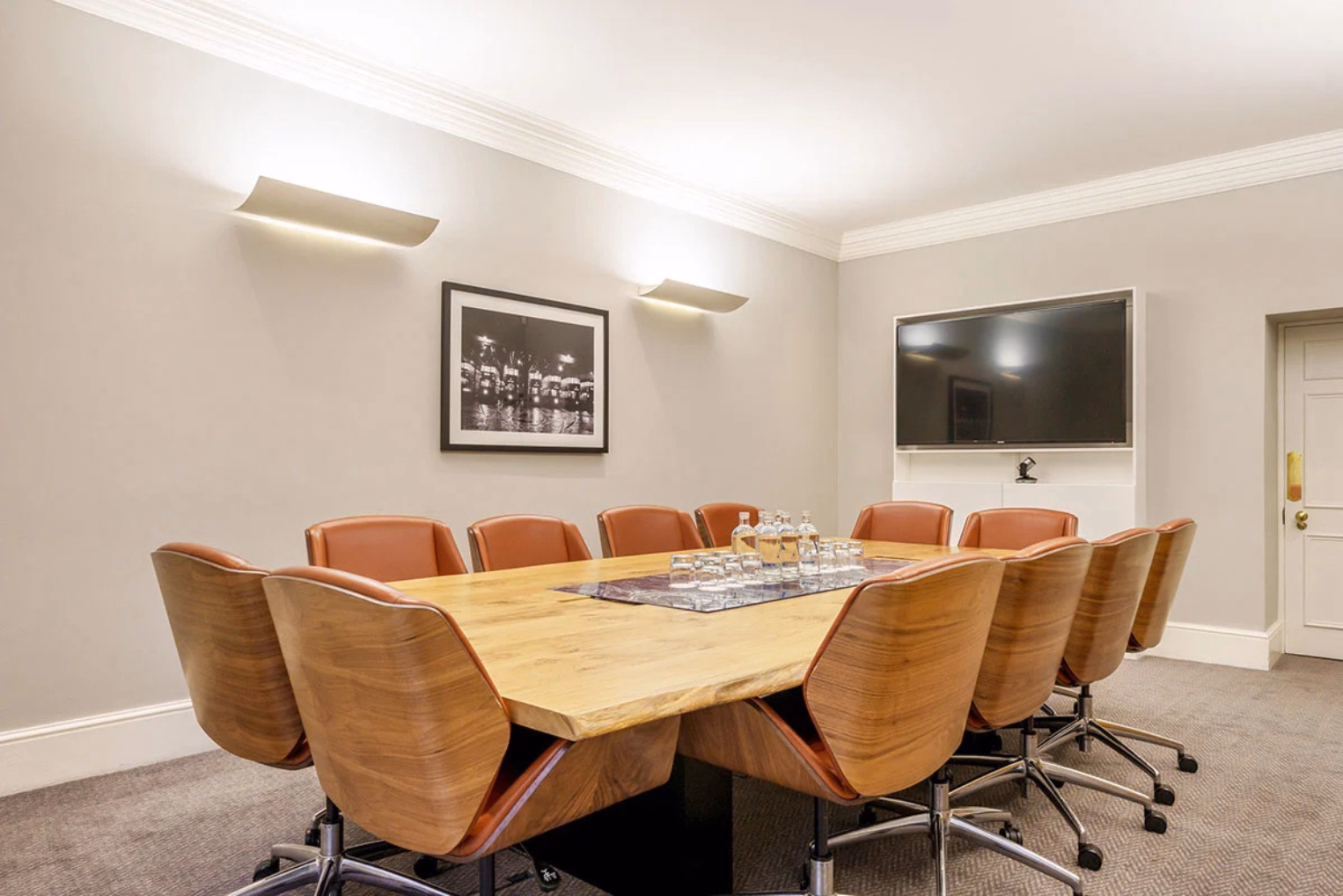 Brown leather chairs surround a large wooden table in a meeting room with a wall-mounted TV.