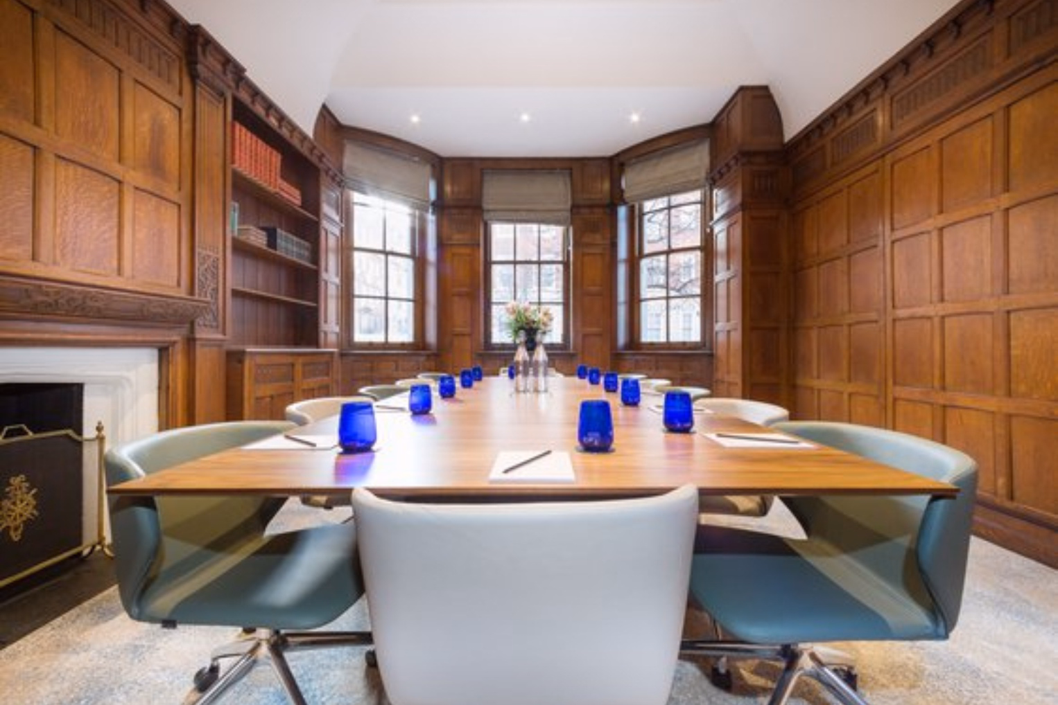 Greyish-blue and white chairs around a long wooden table with blue drinking glasses and wood-panelled walls.