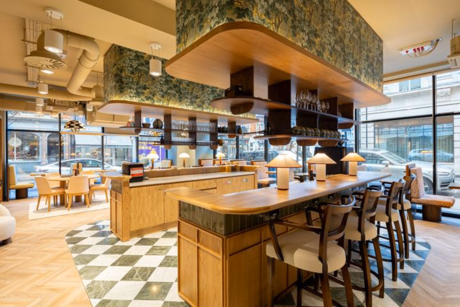 Wooden barstools line a curved wooden breakfast bar topped with small lamps and set on green-and-white checkered floor tile, near large street-facing windows.