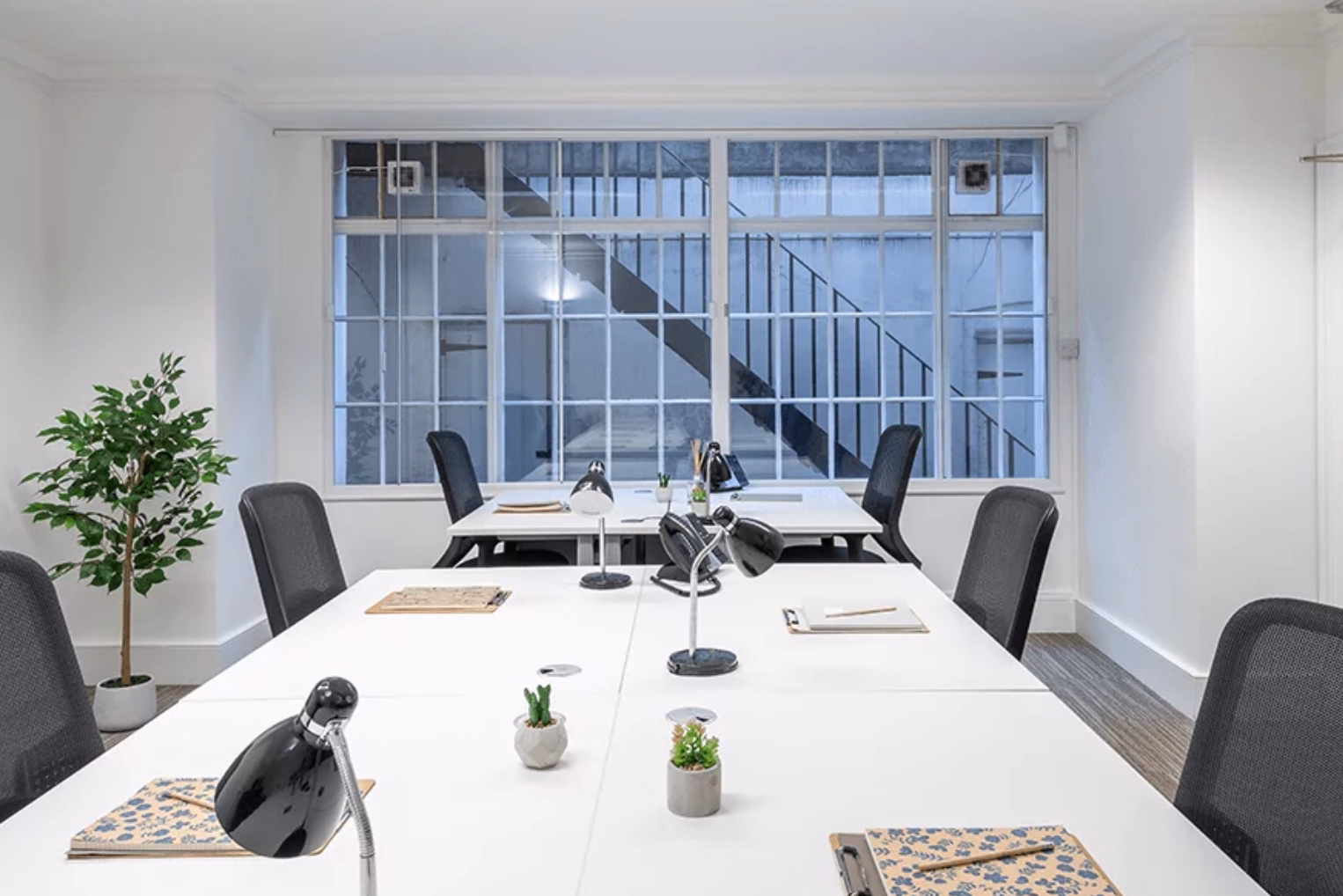 Black mesh chairs surround white work desks equipped with lamps, small potted plants, and notebooks with a window that overlooks a courtyard stairwell.