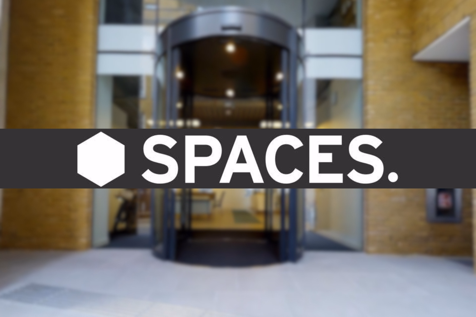 A horizontal banner with the white text "SPACES." overlaid across a blurred image of a building entrance featuring a revolving glass door.
