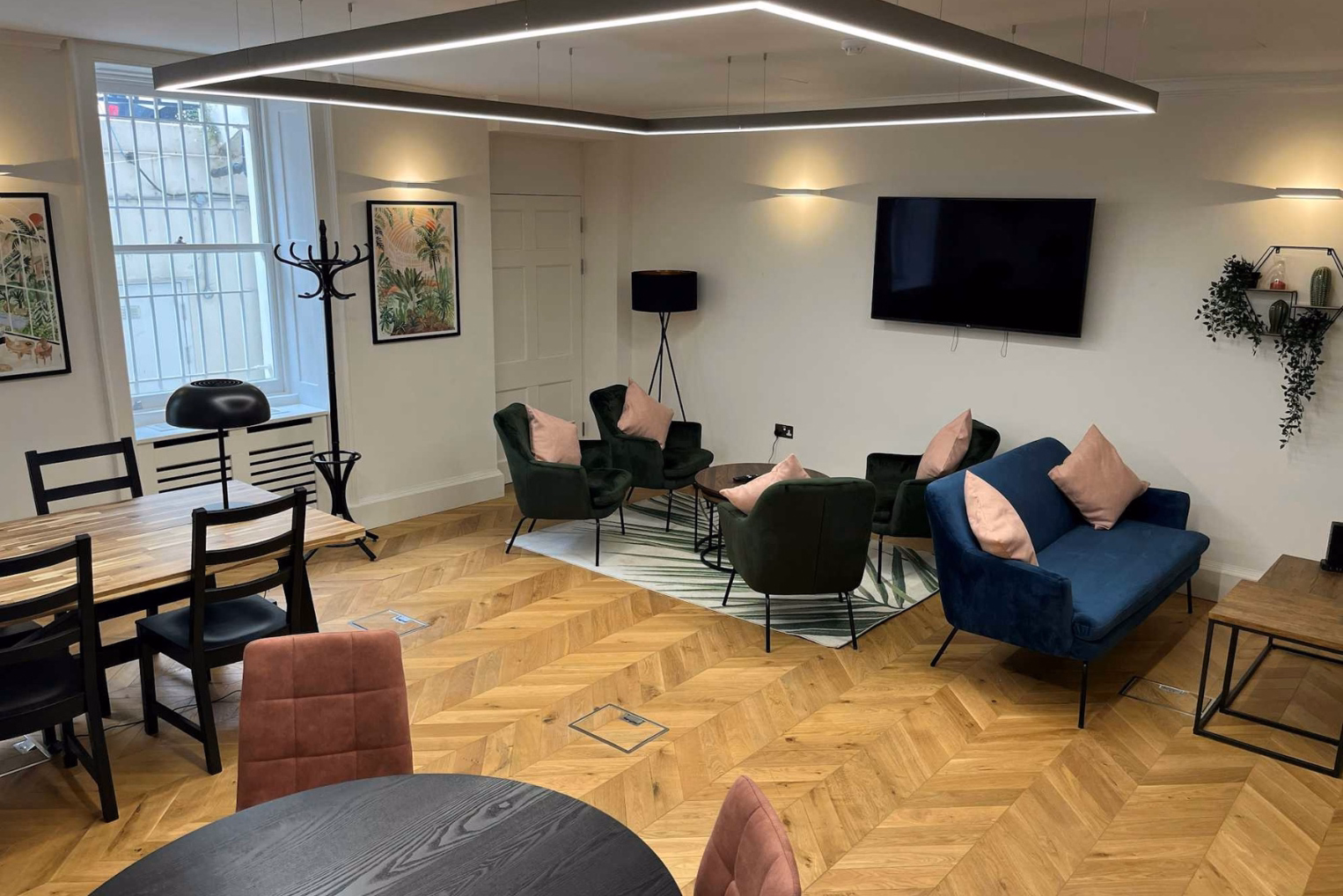 An office lounge and meeting area features green velvet armchairs with pink pillows alongside a blue velvet sofa and a wood-topped dining table with black chairs.
