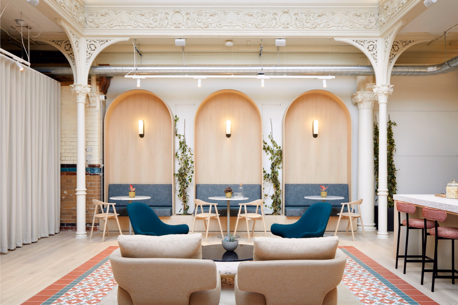 Beige armchairs and dark teal loungers facing three arched wood-panelled seating alcoves with blue cushioned benches, light wooden chairs, and round tables.