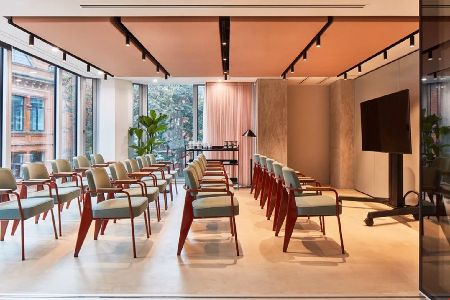 Rows of light green chairs with red frames in a presentation room, facing a large display screen on a rolling stand near floor-to-ceiling windows.
