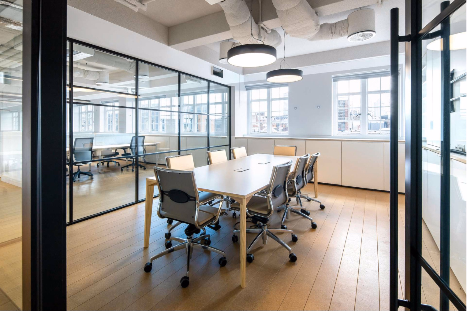 Light grey leather chairs surround a long table in a glass-walled meeting room, which features large windows, two circular pendant lights, and exposed ceiling ductwork.