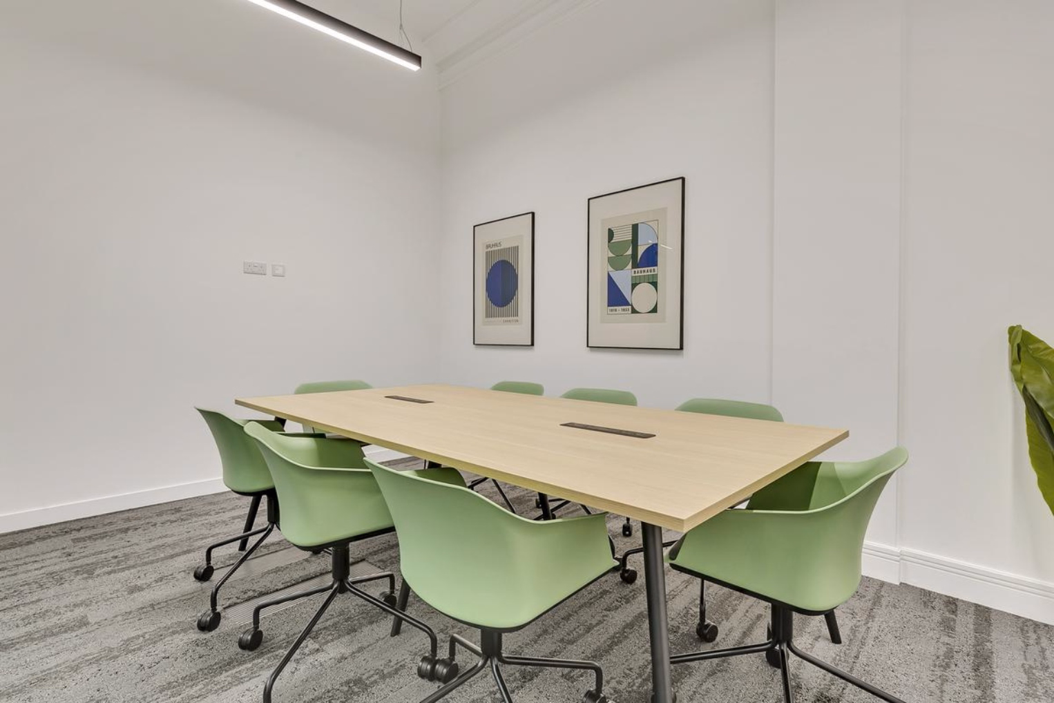 Green swivel chairs surround a light wood rectangular table in a meeting room, complete with grey carpet flooring.