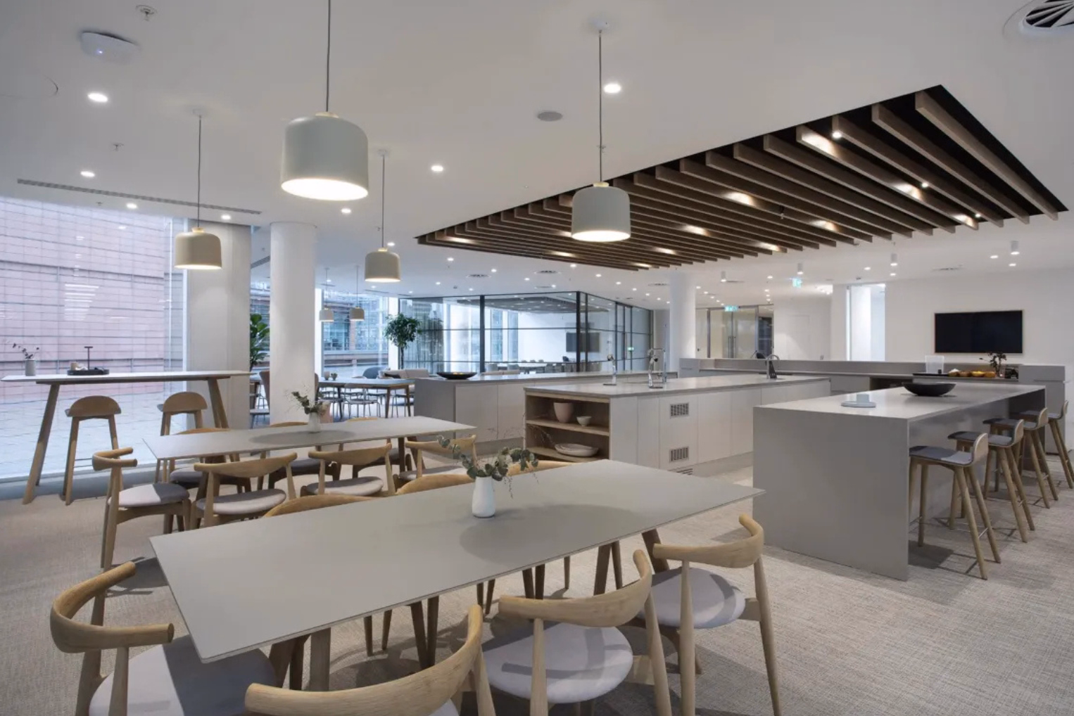 Light wooden dining chairs surround long grey tables in a cafeteria and breakout area, which features grey island counters with matching barstools.