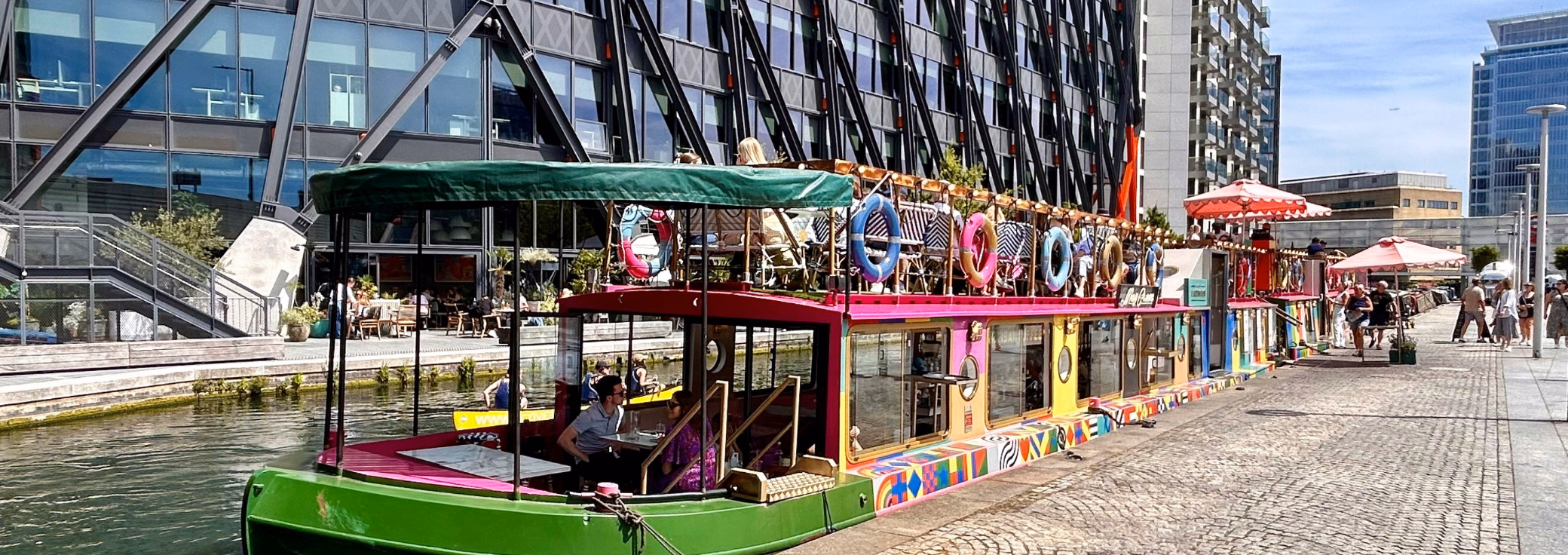 A colourful canal boat filled with outdoor rooftop seating and passengers glides along a waterway next to glass-fronted office buildings under a sunny sky.