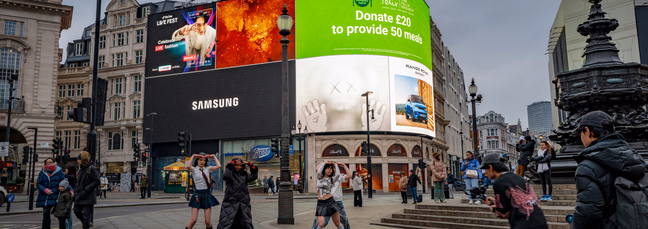 Pedestrians walk and pose for photos in front of a billboard featuring advertisements for Samsung and a charity drive under a grey sky.