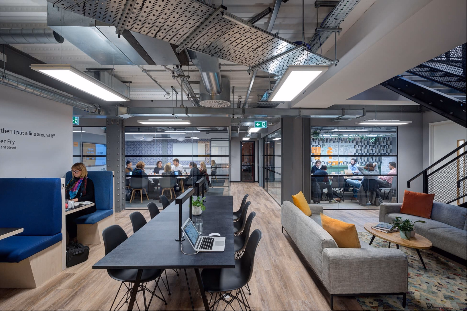 People work in an office and lounge featuring a long black desk, grey sofas with colourful pillows, blue booth seating and glass-walled rooms.