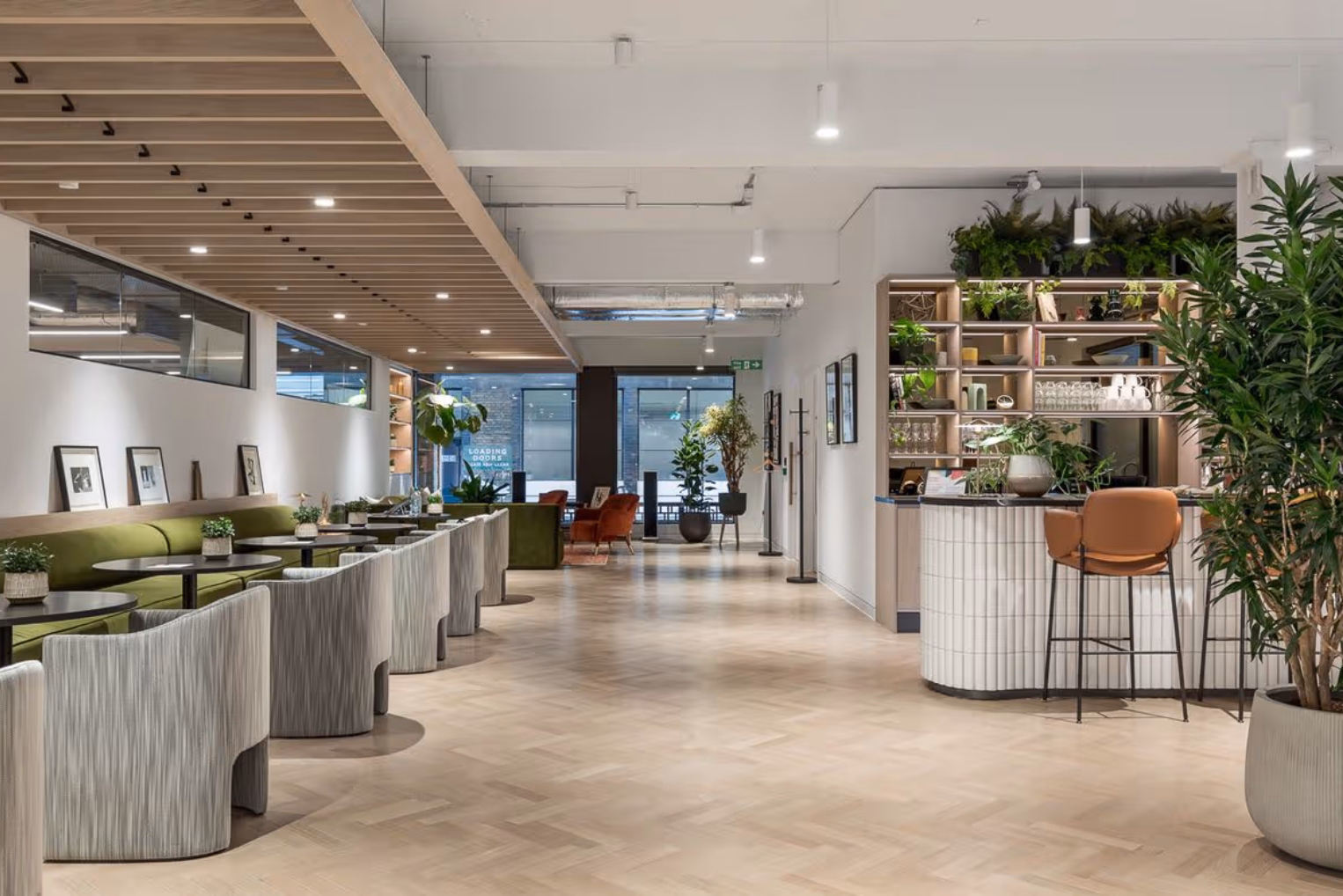 Grey chairs and an olive-green banquette bench along dark circular tables featuring a tiled curved bar counter with leather stools opposite indoor plants.