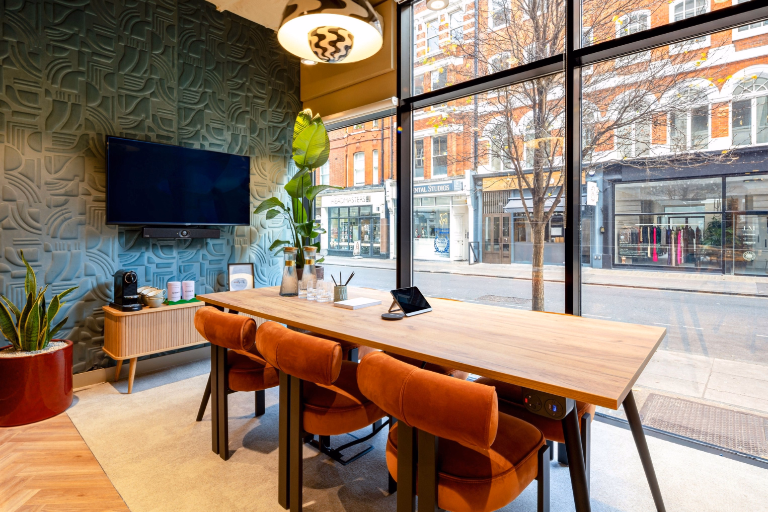Orange chairs surround a wooden table set beside floor-to-ceiling windows looking out onto a street with a wall-mounted TV screen and potted plants.