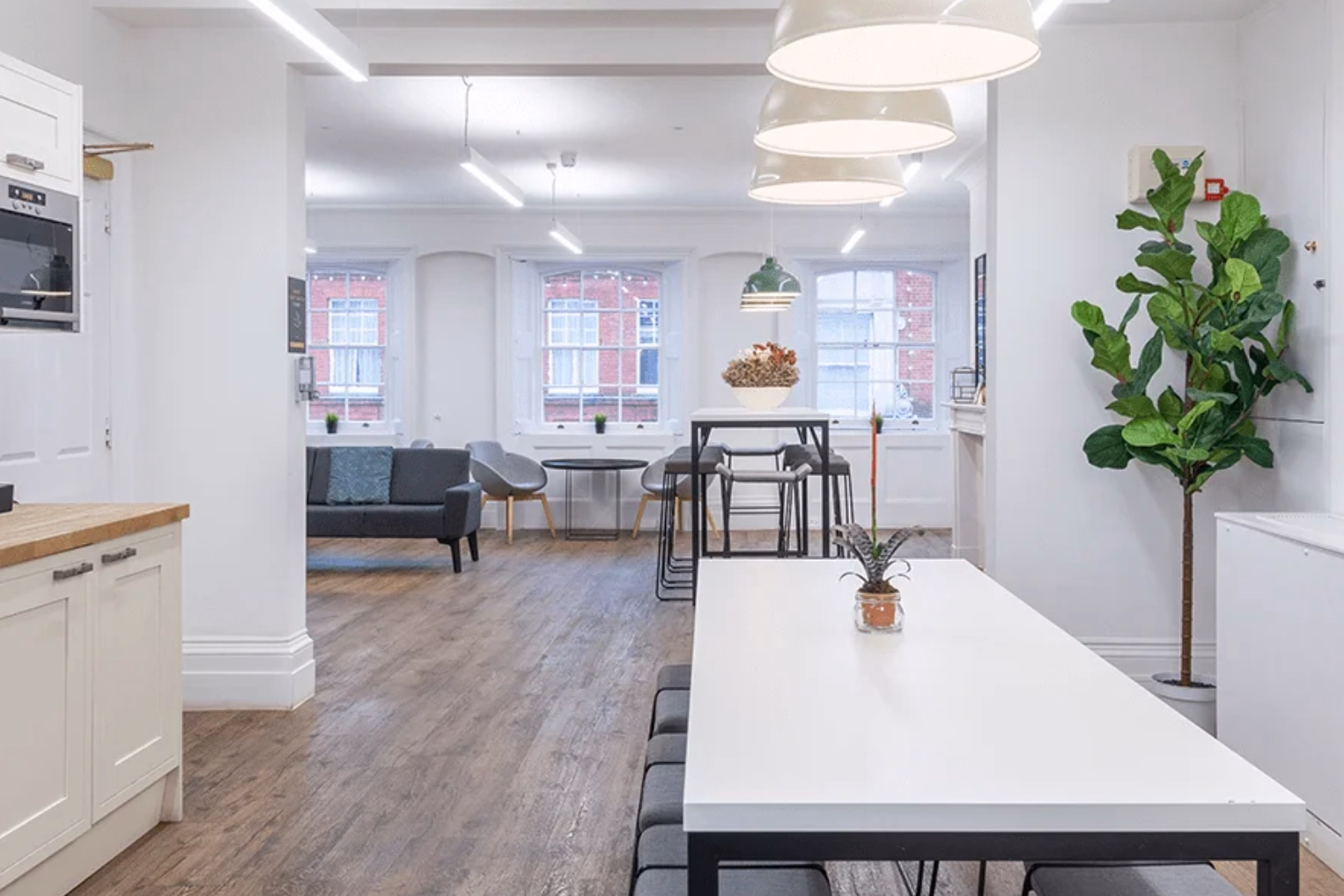 Large pendant lights illuminate a bright open kitchen and break room area featuring a white dining table with grey bench seating.