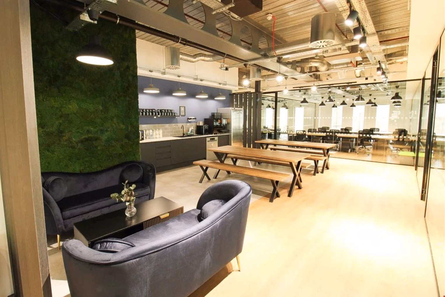 Velvet sofas around a black table alongside wooden picnic-style benches, a kitchenette, exposed ceiling steel beams, and glass-partitioned workspaces.