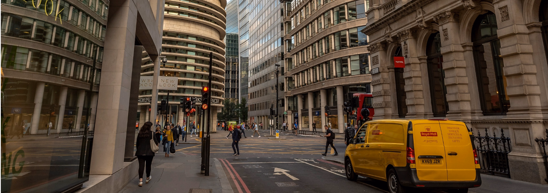 A yellow Royal Mail van is parked along the curb, framed by office buildings, pedestrians at a crosswalk, and red traffic lights.