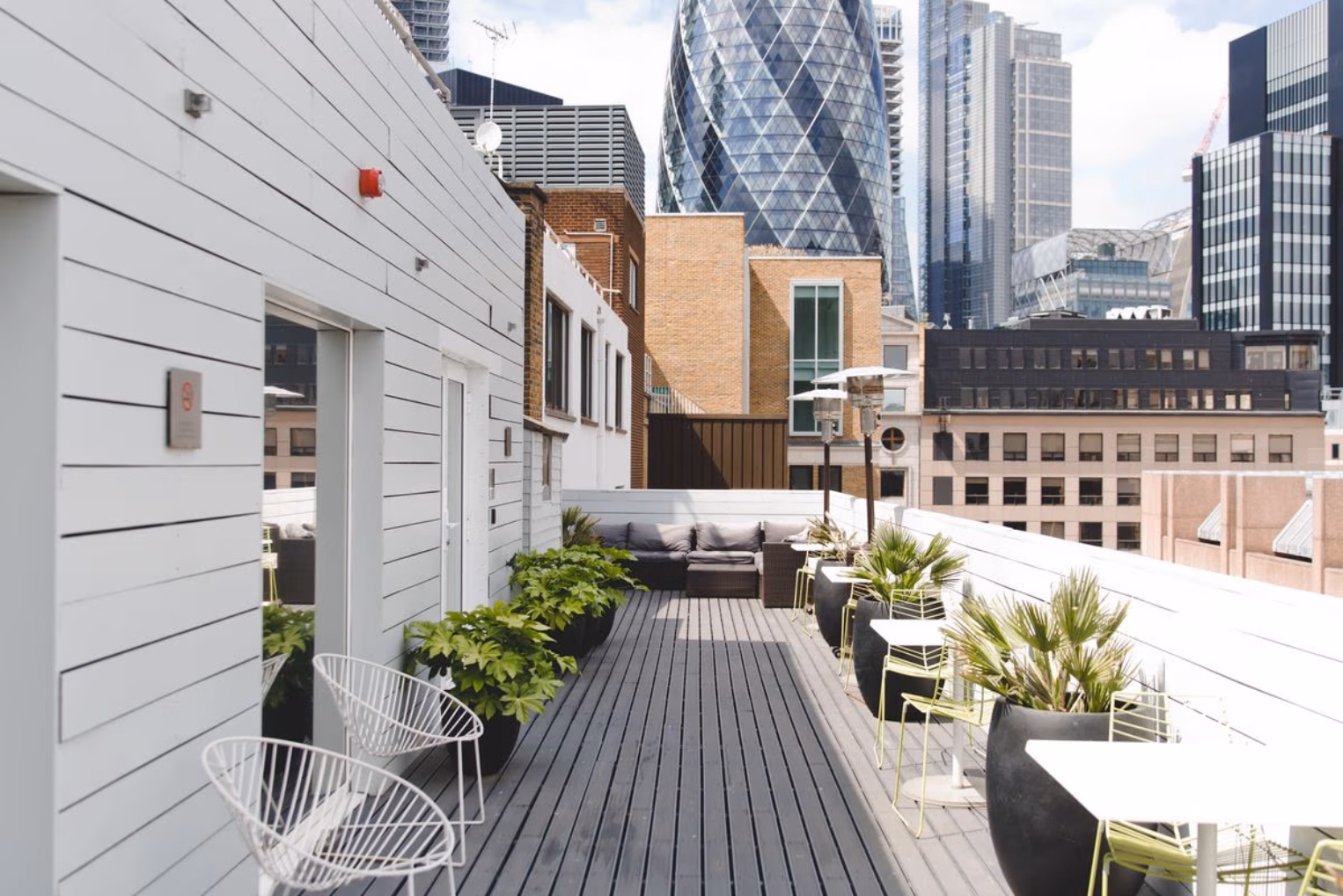 White patio seating and potted plants line an outdoor rooftop deck with scenic views of London city skyscrapers under a bright sky.