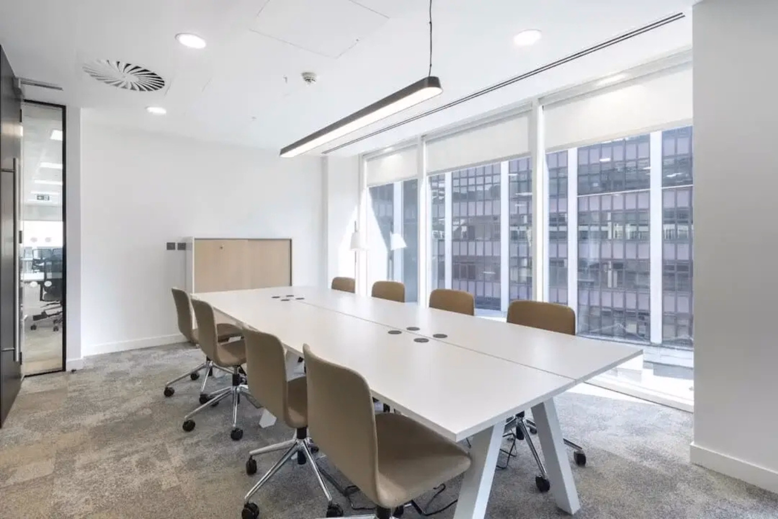 Tan chairs arranged around a white table in a brightly lit meeting room featuring grey carpeting, white walls, and floor-to-ceiling windows fitted with roller blinds.