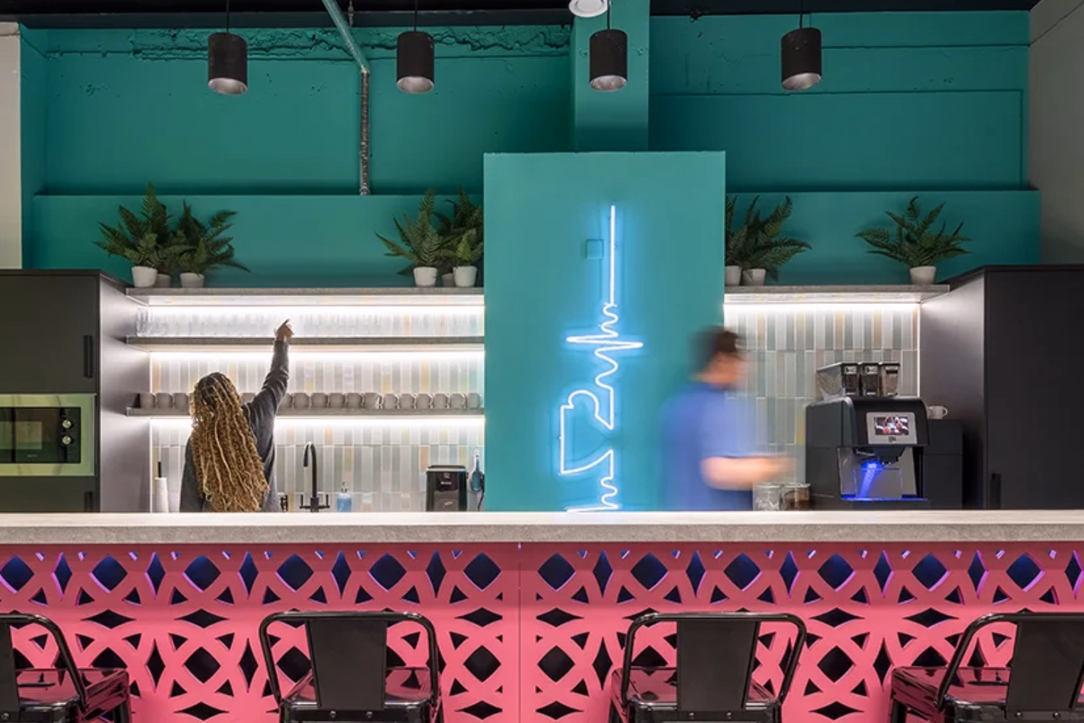 Black metal barstools near a bright pink patterned front panel, teal walls, illuminated shelving, potted plants, and a glowing sign on a central pillar.