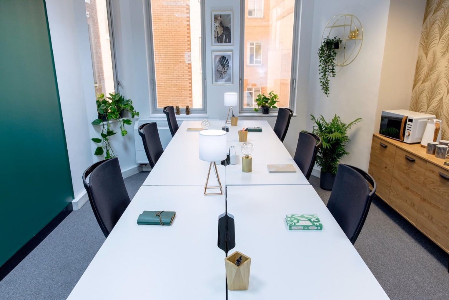 Black chairs along a white table with desk lamps and office accessories, set in an office featuring green walls, light wood cabinets, and potted plants near windows.