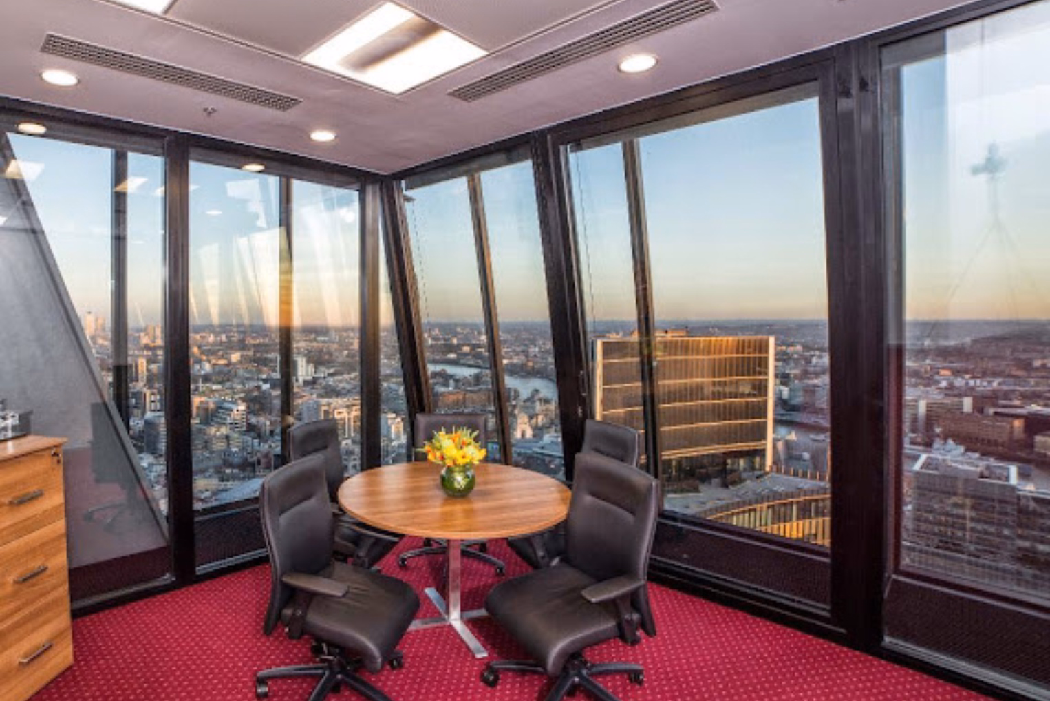 Black leather chairs surround a round wooden table topped with flowers and floor-to-ceiling windows offering views of London's skyline and the River Thames.