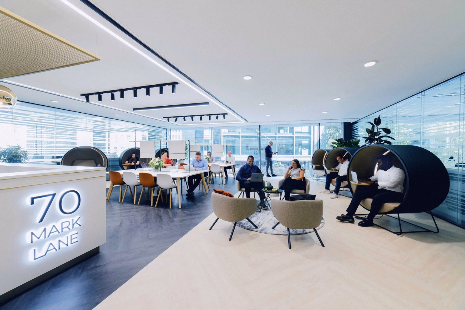 People work in a lounge featuring circular pods, armchairs, communal worktables, and floor-to-ceiling windows, by a reception counter reading "70 MARK LANE."