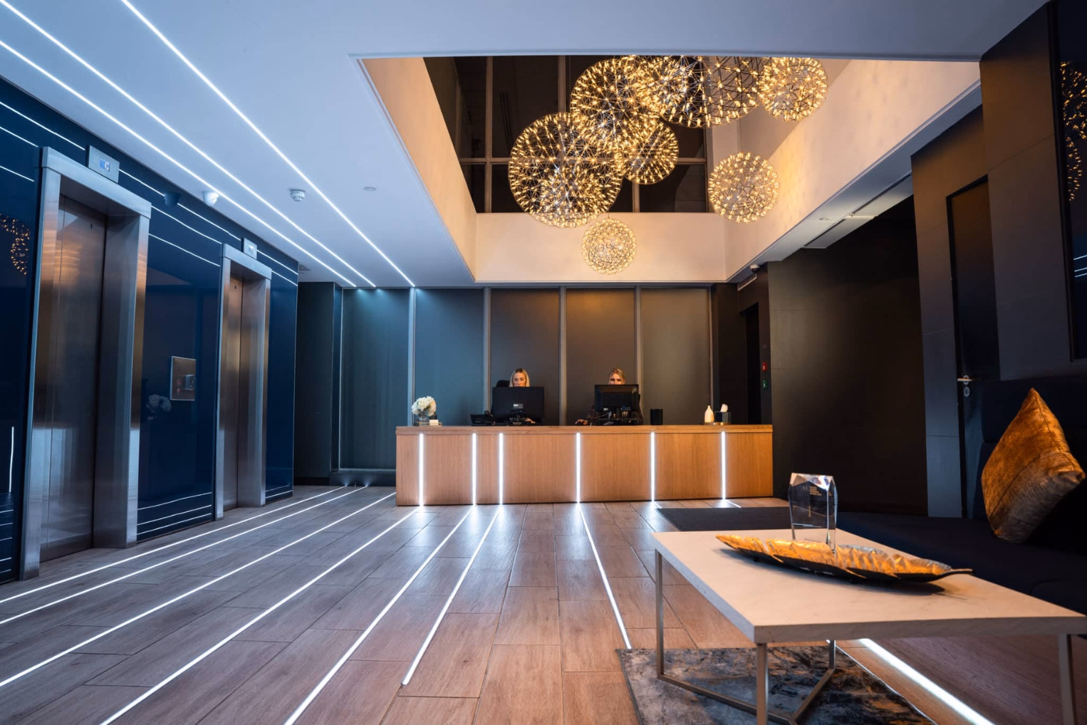 Wire-frame orb chandeliers hang over a long wooden reception desk in a waiting area, highlighted by lighting running continuously along the ceiling, walls, and wood floor.
