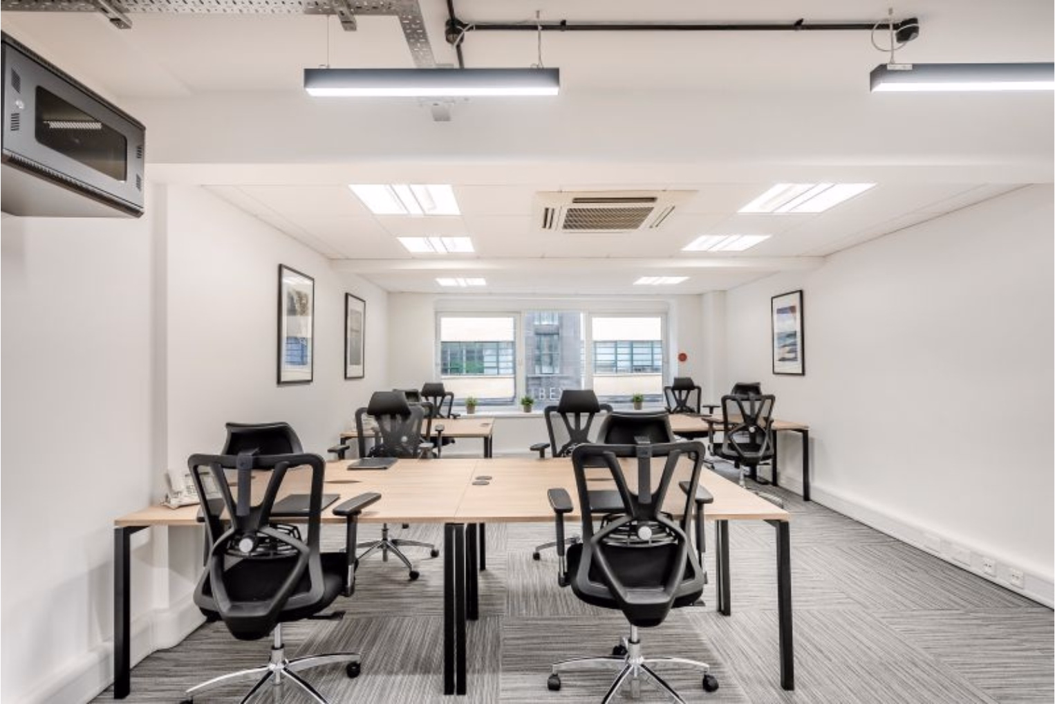 Black chairs at light wood workstations in a brightly lit office space featuring grey striped carpet, white walls with framed art, and recessed ceiling panels.