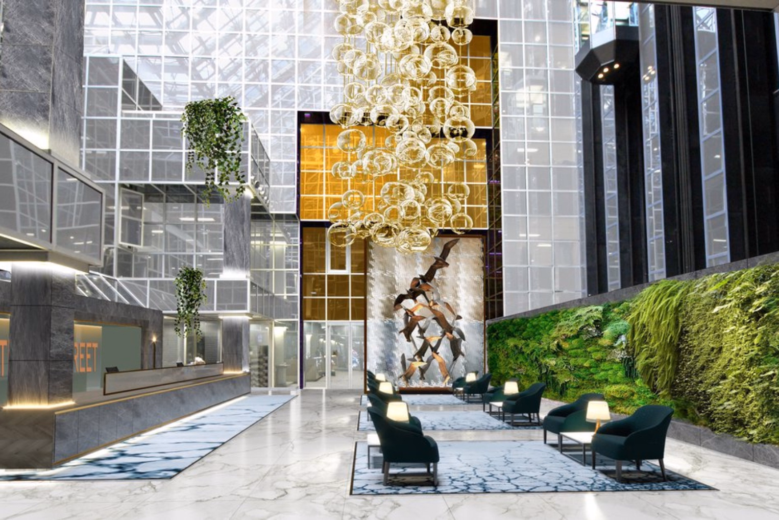 A cascading golden orb chandelier hangs in a glass atrium featuring a plant wall, dark green lounge seating, marble floors, and a reception counter.