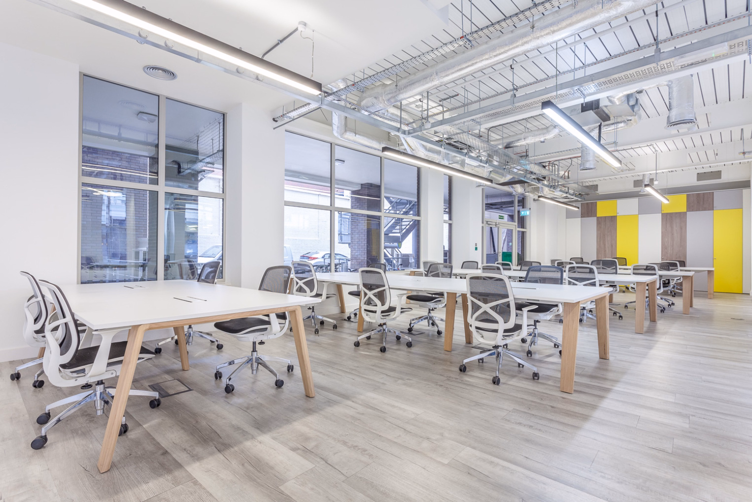 White chairs at white desks in an open-plan office, featuring large windows, exposed ceiling ductwork, suspended lighting, and yellow and grey accent panels.