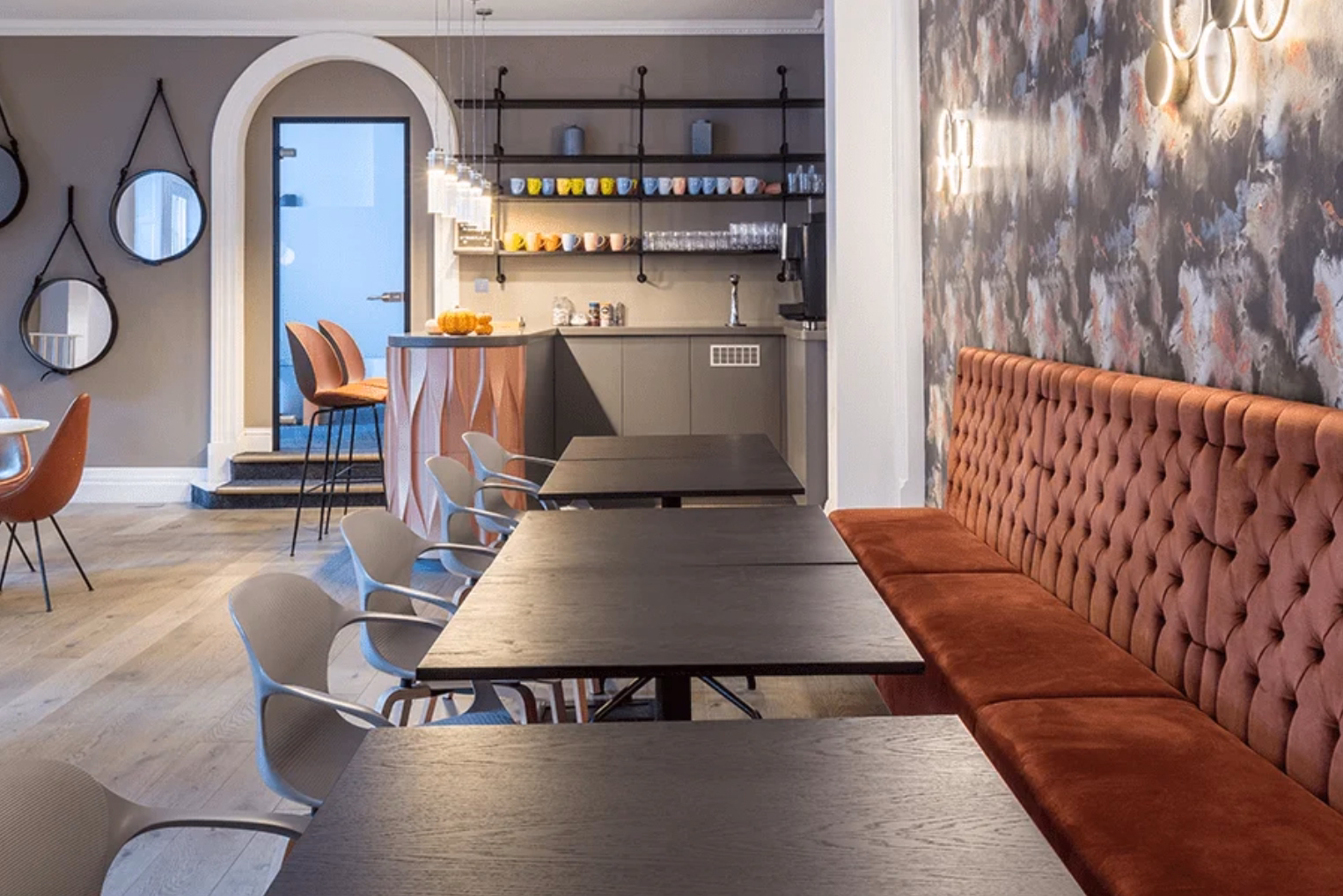 An orange velvet banquette paired with dark wooden tables and grey chairs in a lounge, featuring wall-mounted mirrors and an arched doorway leading to a coffee bar.