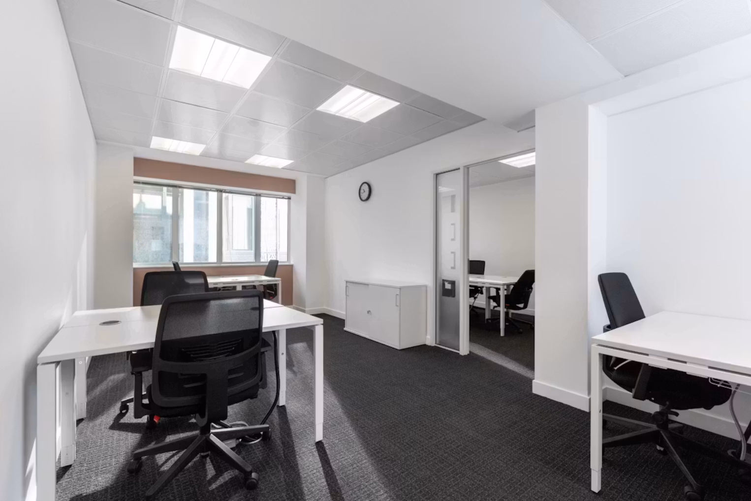 Black chairs with white desks on dark grey carpeting, white walls, ceiling lights, and a glass-partitioned private office room in the background.
