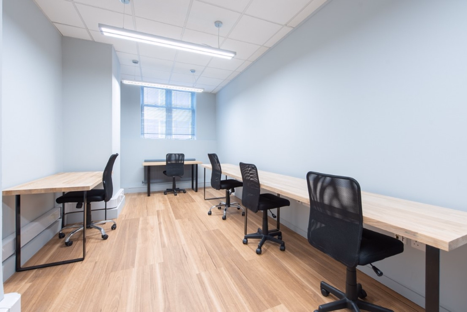 Black chairs along light wooden workstations in a narrow office room featuring grey walls, hanging light fixtures, and a window with blinds at the far end.