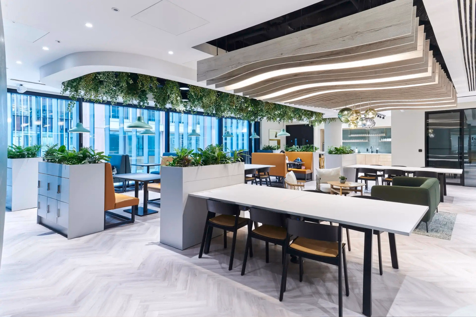 An open-plan office lounge featuring white communal tables, built-in planter dividers, colourful booth seating, and hanging greenery.
