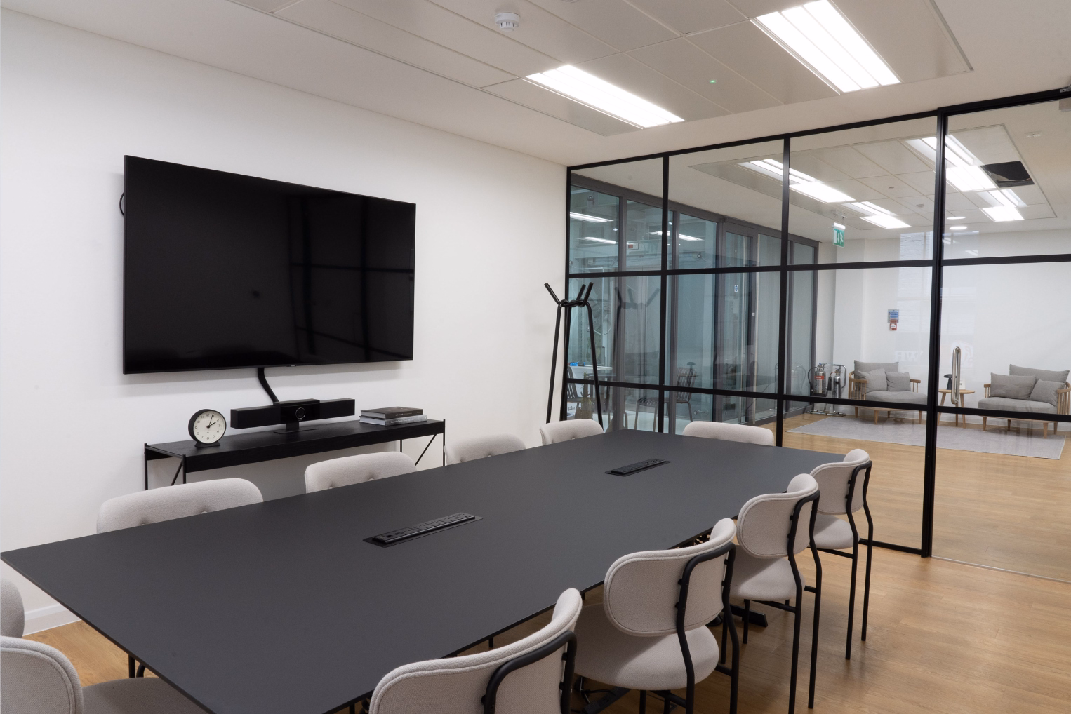Light grey chairs surround a matte black table in a meeting room, featuring a wall-mounted display screen and black grid glass partitions.