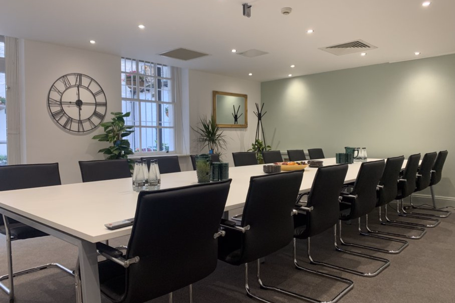 Black leather chairs surround a white table in a boardroom, featuring a green accent wall, a wall clock, potted plants, and ceiling spotlighting.