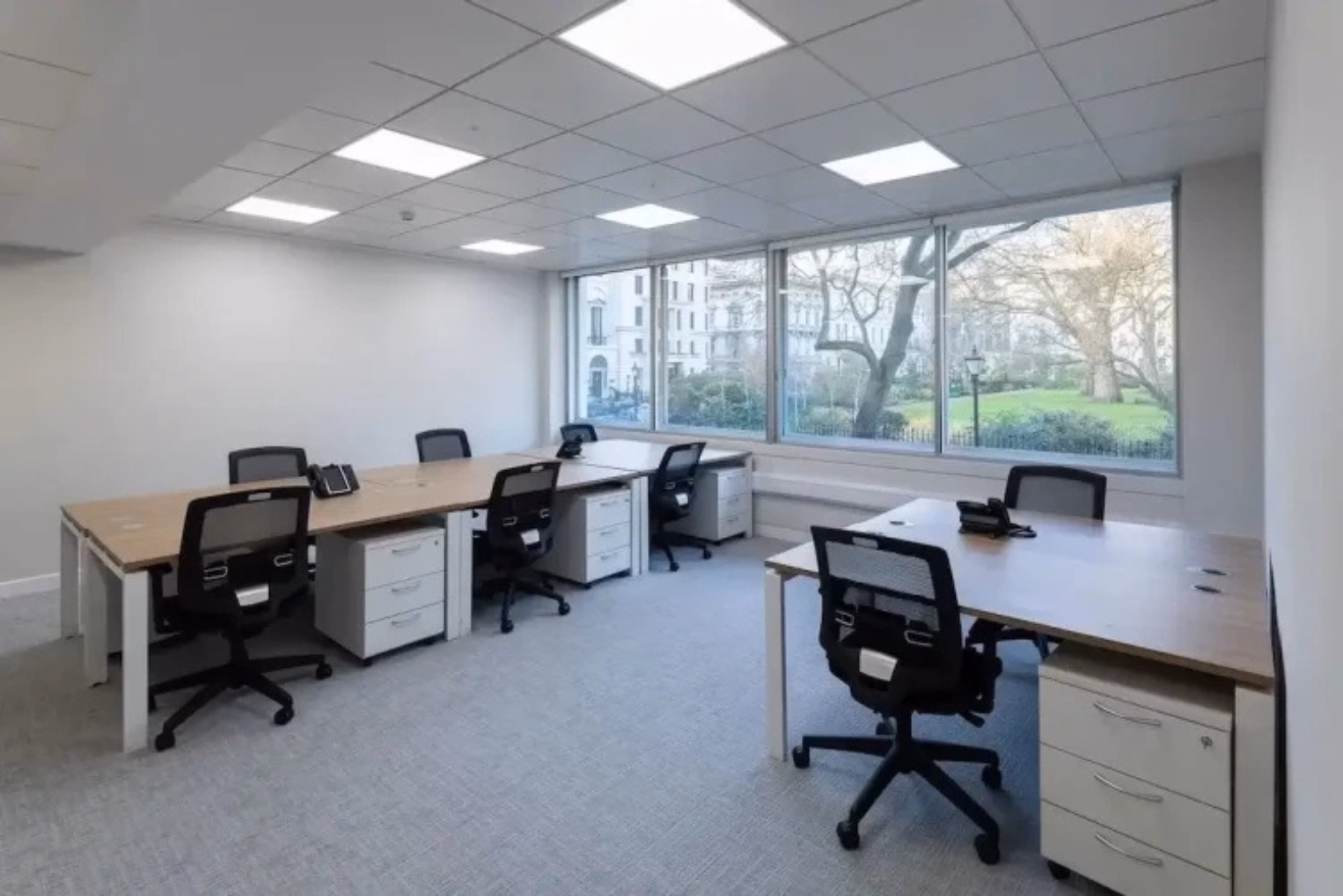 Black chairs are paired with light wood desk pods with under-desk white drawers in an office, featuring white walls, ceiling panels, and expansive windows.